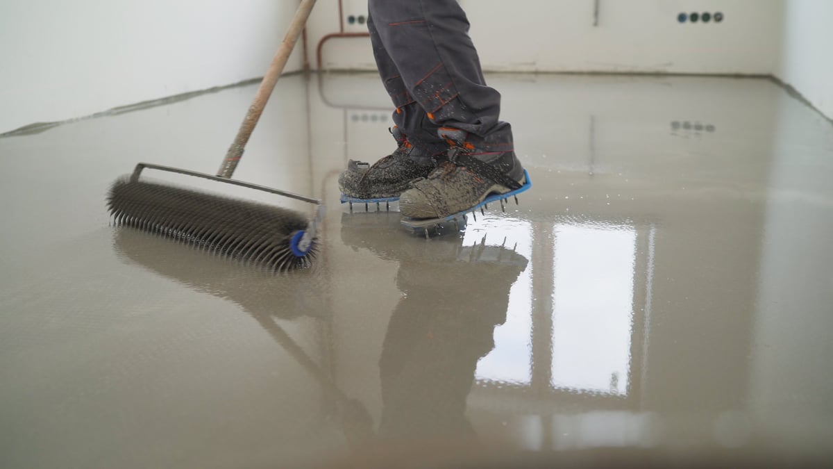 Worker in spiked safety footwear using a roller to spread self-levelling epoxy resin across a reflective grey floor