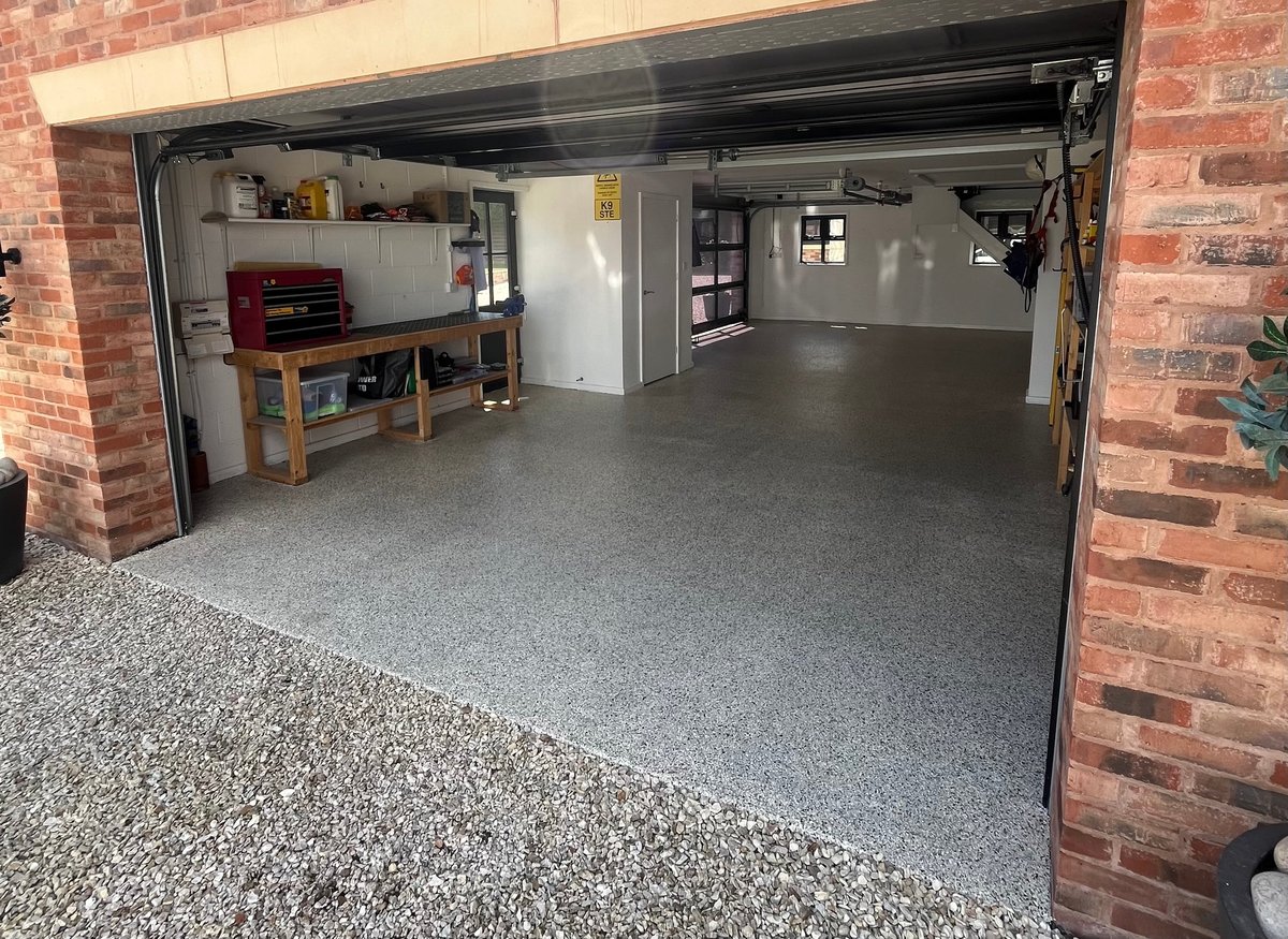 Open garage door showing empty concrete floor with brick walls, shelving units on left side, and exposed ceiling beams