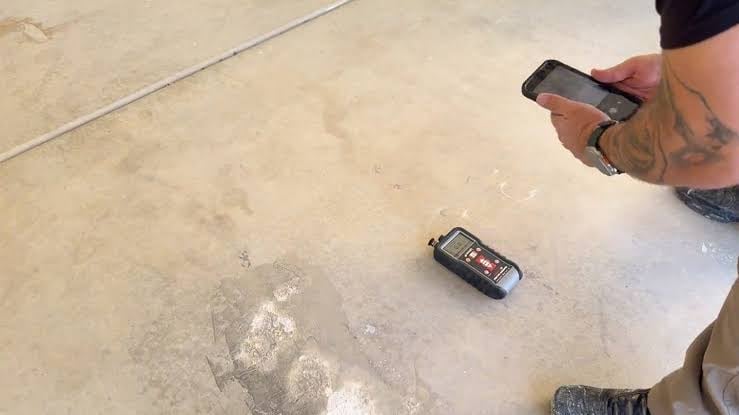 Digital moisture meter placed on a concrete floor with the installer logging readings on a smartphone app during moisture assessment