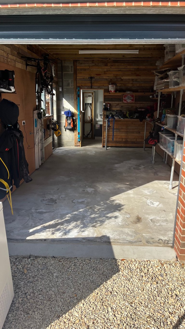 Interior of a garage with wooden walls, bikes and tools hanging on left side, shelving on right, concrete floor, and open doorway leading inside