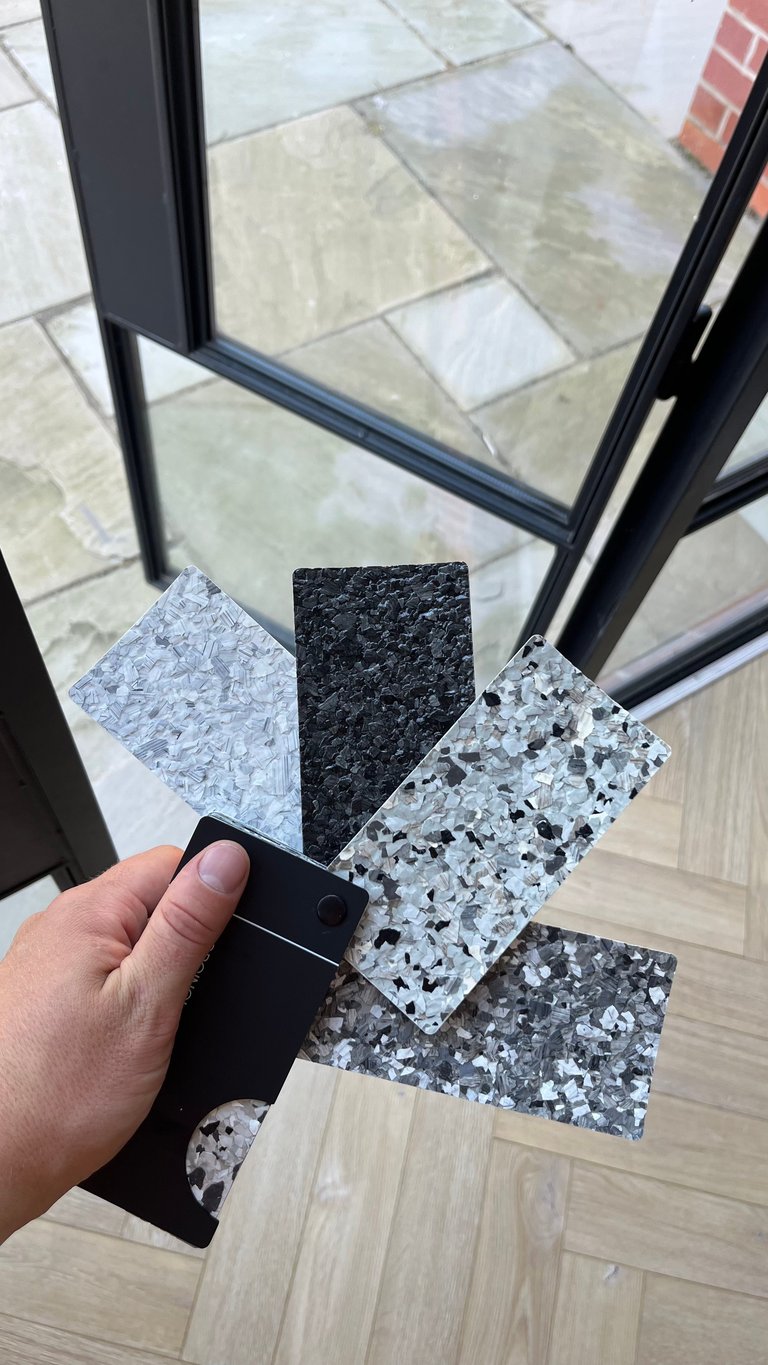 Person holding material samples showing various countertop and flooring options in black, white speckled, and light wood finishes